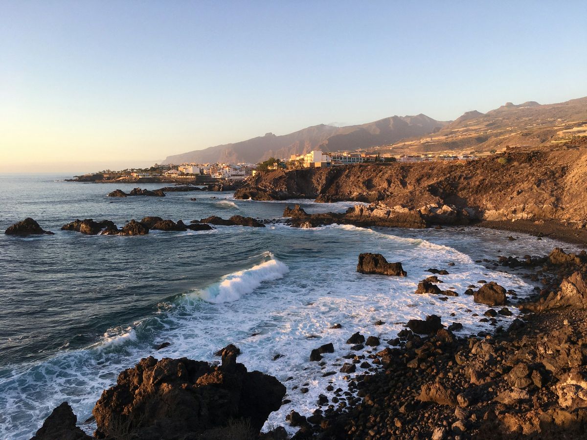 Waves breaking on black volcanic rocks on Tenerife's west coast, with a white town and mountains behind at sunset