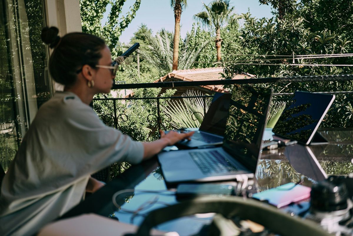 A woman working at a laptop on a terrace surrounded by palm trees